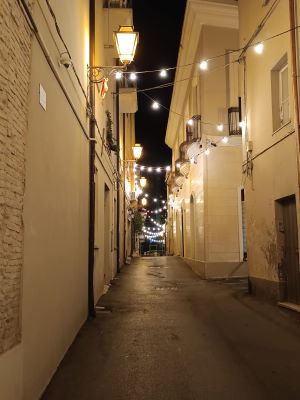Il garante relazionale - Vicolo cittadino notturno illuminato da catene di luci sospese e lampioni a parete, con pavimentazione in asfalto ed edifici storici ai lati.