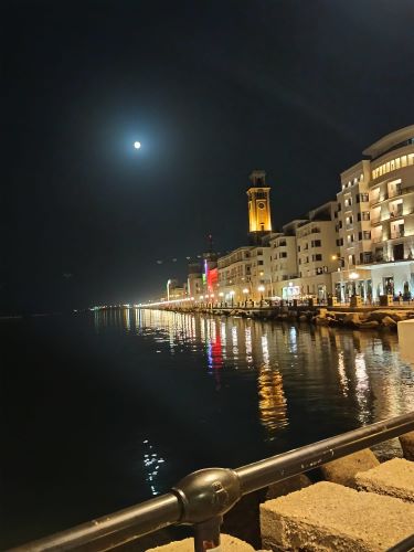 Diplomazia dei Sentimenti - Lungomare di Bari di notte con la luna piena e la torre dell'orologio illuminate.