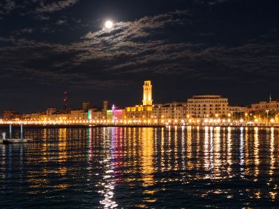 Panorama notturno del lungomare di Bari illuminato, con il riflesso delle luci sull'acqua del mare e la luna piena in cielo, simbolo della visione relazionale e comunitaria della "Diplomazia dei Sentimenti".